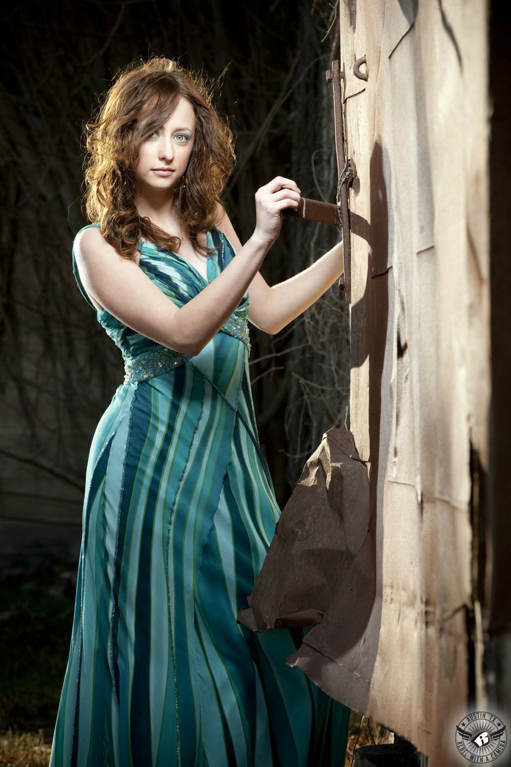 curvy red curly haired, green eyed model with a aquamarine striped full length dress with straps leans against rusted metal door with ripped metal